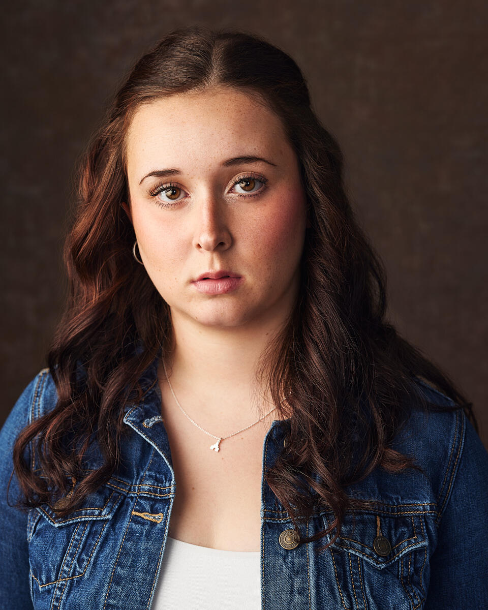A character style headshot of a woman wearing a denim jacket shot on a brown canvas background.